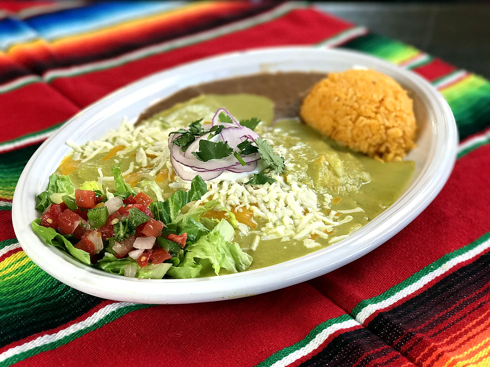 Enchiladas verdes on a white oval plate, with shredded cheese, red onion, rice, beans, lettuce, and pico de gallo
